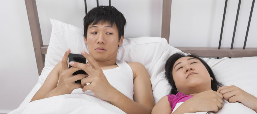 infidelity private investigator photo of a man sneaking a look at phone while in bed with wife
